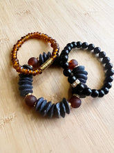Load image into Gallery viewer, The edgy combo of black and amber creates a unique and gorgeous stack. Features a black ashanti glass bracelet with with recycled glass beads, an amber glass bracelet with brushed gold accents and an onyx bracelet with small gold and crystal accent bead All bracelets measure 7 inches but can be customized upon request