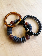 Load image into Gallery viewer, The edgy combo of black and amber creates a unique and gorgeous stack.  Features a black ashanti glass bracelet with with recycled glass beads, an amber glass bracelet with brushed gold accents and an onyx bracelet with small gold and crystal accent bead  All bracelets measure 7 inches but can be customized upon request
