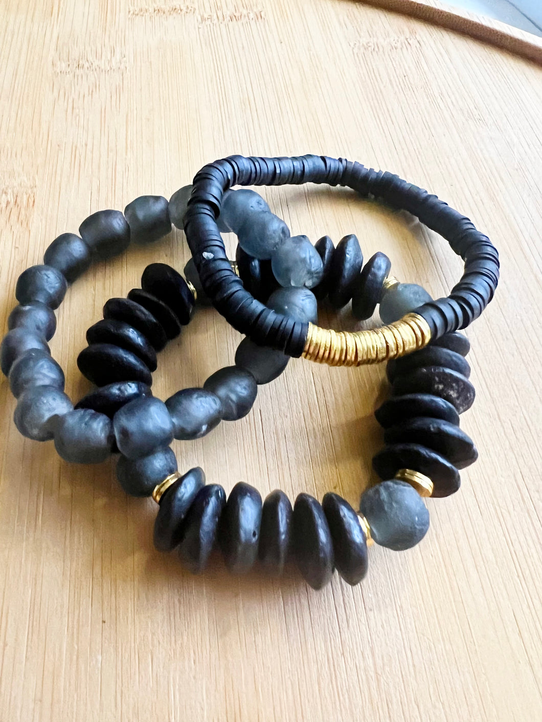 A classic black stack featuring a black ashanti glass bracelet with with recycled glass beads, a black polymer clay bracelet with brushed gold accents, and a smokey black recycled glass bracelet  All bracelets measure 7 inches but can be customized upon request